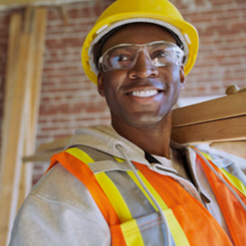 um homem sorrindo com capacete amarelo e segurando madeiras sobre um dos ombros
